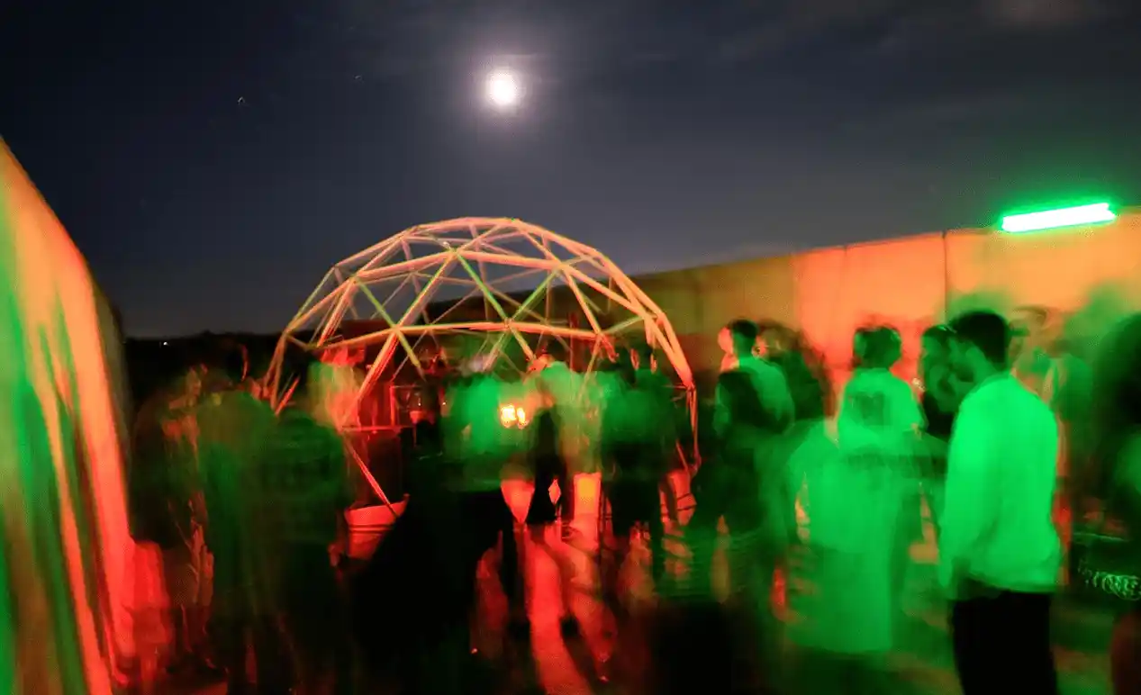 Una scena di festa notturna all'aperto sotto la luce della luna piena, con persone sfocate in movimento illuminate da luci verdi. Un'illuminazione rosso-verde prevale sulla scena, con una struttura a cupola geodetica al centro dove si radunano le persone.