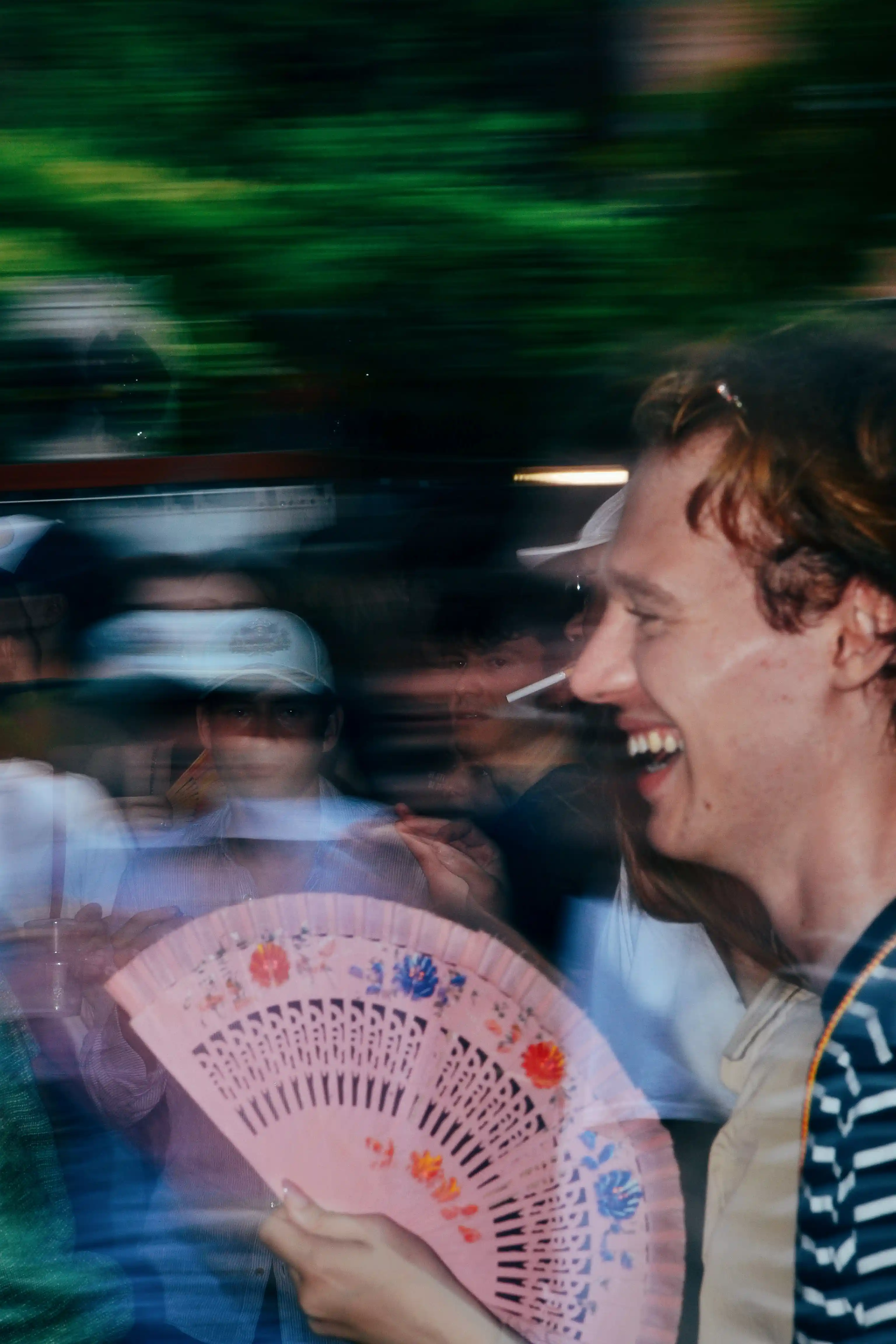 Foto di un uomo giovane, sorridente, che tiene un ventaglio rosa aperto. L'immagine è catturata con un effetto di movimento, mostrando sfocature di persone e luci di sfondo.