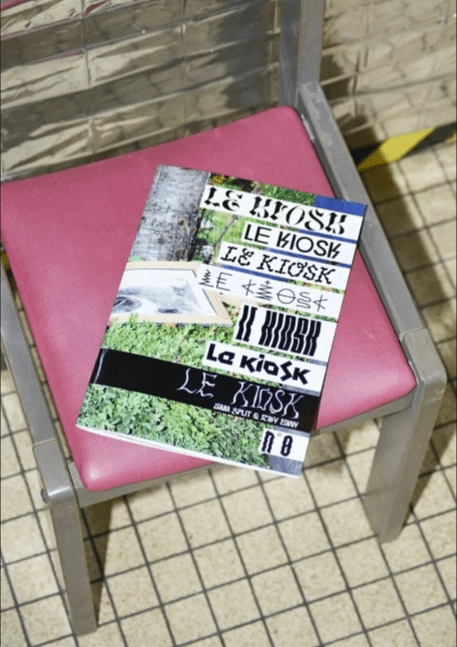 Una rivista intitolata 'LE KIOSK' posata su una sedia con seduta rosa in una stanza con piastrelle bianche. La copertina della rivista presenta una disposizione collage di testi e immagini, con il nome 'LE KIOSK' ripetuto in diversi stili tipografici.